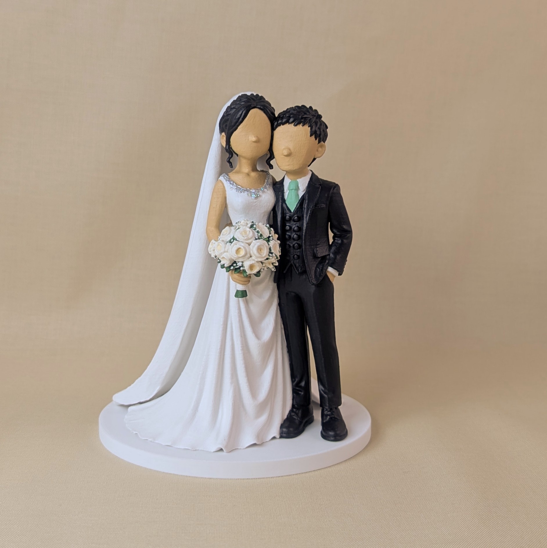 Custom wedding cake topper of a bride in a beaded-neckline gown and cathedral veil holding white roses, beside a groom in a black three-piece suit and mint green tie