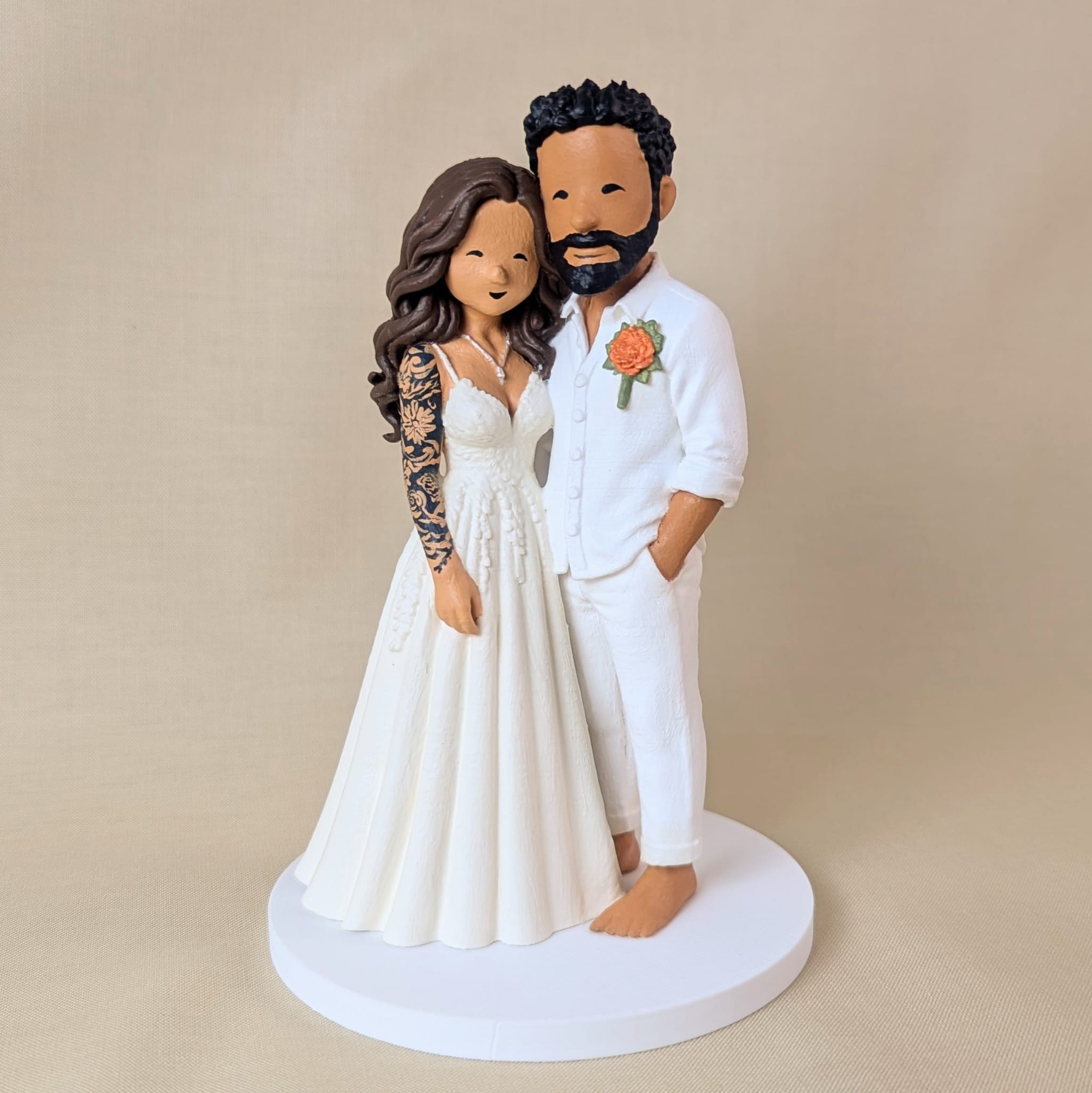 Custom wedding cake topper of a bride in an ivory lace gown with a detailed floral sleeve tattoo, beside a barefoot groom in a white linen shirt and trousers with a marigold boutonniere