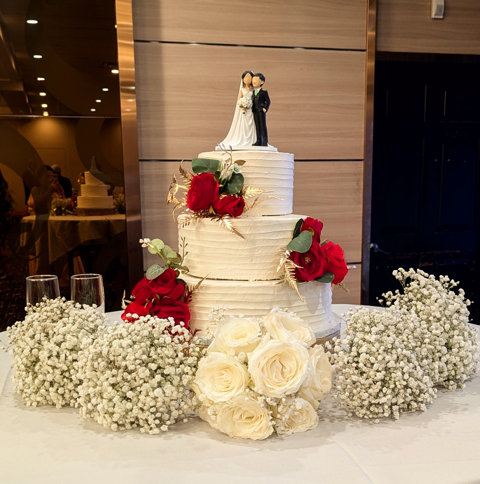 Custom bride and groom cake topper on a three-tier white buttercream wedding cake decorated with red roses and gold foliage